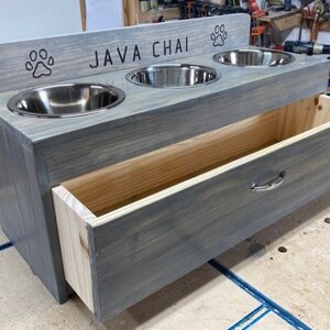 Pu&ograve; includere: Una mangiatoia per animali domestici in legno grigio con tre ciotole in acciaio inossidabile e un cassetto. La parte superiore reca la scritta "JAVA CHAI" e impronte di zampe. Il cassetto ha una maniglia argentata.