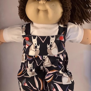 May include: A doll dressed in a white t-shirt and a dark blue jumpsuit. The jumpsuit is patterned with kangaroos and joeys, alongside stylized floral and leaf designs in grey, pink, and orange. The doll has brown hair.