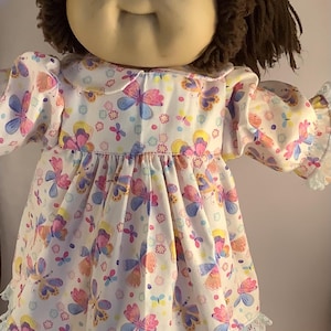 May include: A vintage doll with a round face, brown hair, and a white dress decorated with colourful butterflies. The dress has a ruffled collar, sleeves, and hem, with blue lace trim. The doll's eyes are brown.
