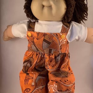 May include: A Cabbage Patch Kid doll with brown hair wearing a white shirt and orange dungarees with a brown and white pattern of kangaroos and plants.