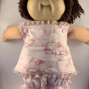 May include: A vintage Cabbage Patch Kid doll with brown hair and a light skin tone. The doll is wearing a pink and white outfit with a Winnie the Pooh print and lace trim. The outfit includes a top and shorts.