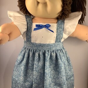 May include: A Cabbage Patch Kid doll wearing a blue and white dress with a white lace trim. The dress has a blue paisley pattern and a blue bow at the neckline.