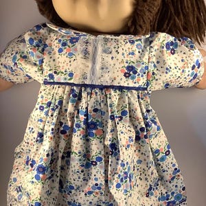 May include: A vintage doll with a round face and brown hair. The doll is dressed in a white dress with a blue floral pattern, blue trim, and white lace detailing. The dress has short sleeves and a Peter Pan collar.