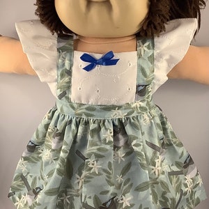 May include: A Cabbage Patch Kid doll wearing a white and blue dress with a floral pattern and blue bow. The dress has a white bodice with a lace trim and a blue skirt with a floral pattern featuring birds.