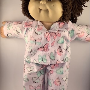 May include: A Cabbage Patch Kid doll wearing a lavender pyjama set with a butterfly print. The pyjamas have long sleeves and long trousers. The doll has brown yarn hair and a pink button on the front of the shirt.