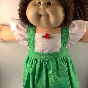 May include: A Cabbage Patch Kid doll wearing a green and white dress with a red rose accent. The doll has brown yarn hair and a white lace trim on the dress.