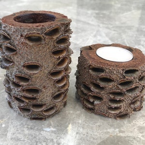 May include: Two cylindrical candle holders made from a textured, brown material. One is taller and empty, while the other is shorter and holds a white tea light candle. The holders have a unique, organic appearance.