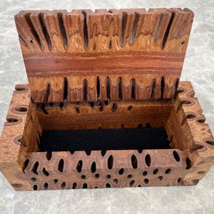 May include: A wooden box with a hinged lid, crafted from reddish-brown wood. The box features a distinctive pattern of holes and grooves. The interior is lined with black fabric. The lid is slightly open, showing the inside.