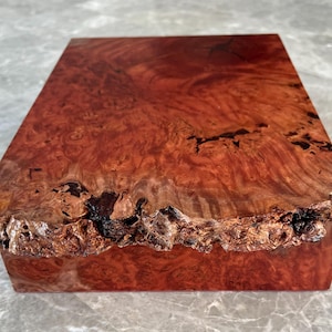 May include: A rectangular wooden box with a rich, reddish-brown burl wood grain. The top surface displays a swirling pattern, while the edges have a rough, natural bark texture. The box is likely used for storage or display.