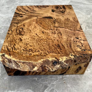 May include: A rectangular wooden block with a natural, unfinished edge. The wood features a complex grain pattern with shades of brown, tan, and black. The block's surface is smooth, showcasing the intricate details of the wood's texture.