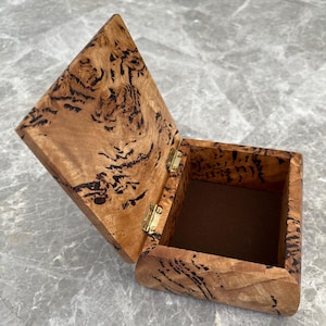 May include: An open wooden box with a hinged lid. The box is made of light brown wood with dark brown, swirling patterns. The interior is lined with a brown fabric. The box is rectangular with a diamond-shaped lid and brass hinges.