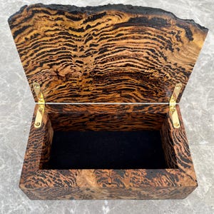 May include: Open wooden box with a hinged lid. The box is made of wood with a unique grain pattern in shades of brown and black. The interior is lined with black fabric, and the hinges are gold-colored.