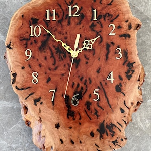 May include: A handcrafted wall clock made from a single piece of wood. The clock has a natural, irregular shape with a warm, reddish-brown hue and dark accents. Gold-coloured hands and numerals complete the design, adding a touch of elegance to this rustic home decor item.