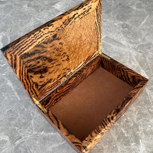 May include: An open wooden box with a hinged lid. The box features a striking tiger stripe pattern in shades of brown and black. The interior is lined with a soft, brown fabric. The box is likely used for storing jewelry or small keepsakes.
