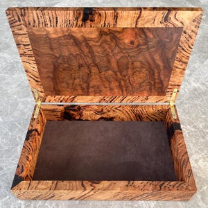 May include: Open wooden box with a hinged lid. The box is made of wood with a unique grain pattern in shades of brown and black. The interior is lined with a brown fabric. The box is open, revealing the interior.