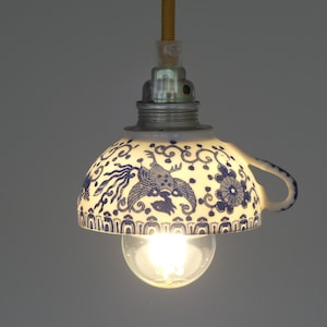 Puede incluir: Una lámpara colgante única hecha de una taza de té. La pantalla blanca de la taza de té presenta patrones florales y de pájaros azules. Una bombilla transparente es visible debajo, suspendida por un cable amarillo. Medidas en cm.