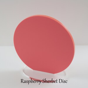 Op de afbeelding: Een ronde, koraal gekleurde schijf van acryl, staande op een heldere acryl basis. De schijf is ongeveer 15 cm in diameter. De tekst "Raspberry Sherbet Disc" is afgedrukt onder de basis.