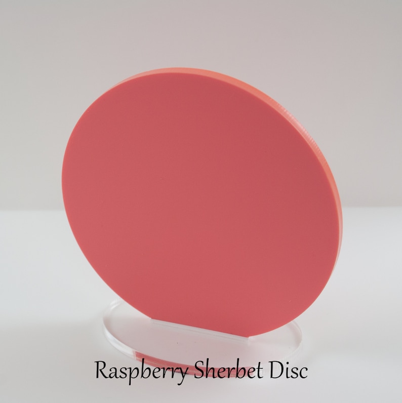 Op de afbeelding: Een ronde, koraal gekleurde schijf van acryl, staande op een heldere acryl basis. De schijf is ongeveer 15 cm in diameter. De tekst "Raspberry Sherbet Disc" is afgedrukt onder de basis.