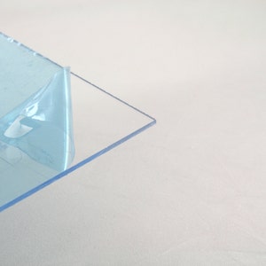 May include: A clear, rectangular sheet of transparent plastic with a protective blue film partially peeled back. The edges of the sheet are clean and sharp, and the overall appearance is smooth and reflective. The background is a plain white surface.