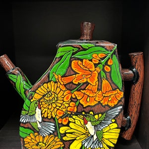 May include: A hand-painted ceramic teapot with a vibrant floral and hummingbird design. The teapot features yellow and orange flowers, green leaves, and two hummingbirds in flight. The handle and spout are brown, and the background is dark.