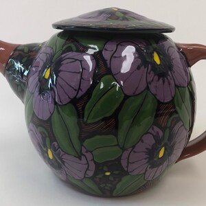May include: A ceramic teapot with a lid, featuring a floral design of purple flowers with yellow centers and green leaves. The spout and handle are brown. The teapot is a rounded shape.
