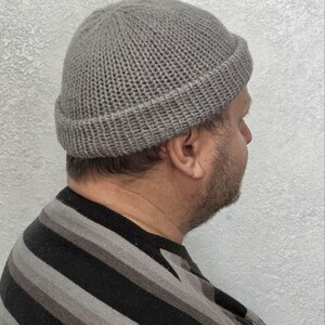 May include: A gray knitted beanie hat with a folded brim. The hat is made of a textured yarn and is worn by a person. The person is wearing a striped black, gray, and white sweater.