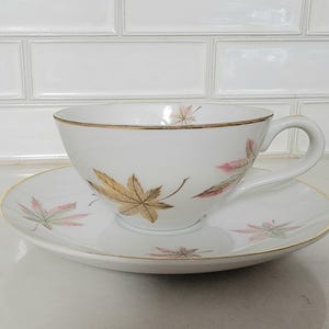May include: White teacup and saucer set with gold trim and autumn leaf designs in gold and pink. The teacup has a curved handle and sits on a matching saucer. A classic tea set for serving tea or coffee.