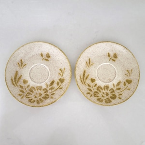 May include: Two white ceramic saucers with a gold floral design. The saucers have a speckled texture.