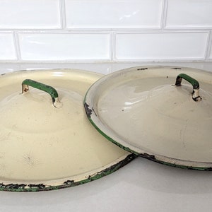May include: Two vintage white enamelware lids with green rims and handles. The lids are slightly worn and have a rustic look.