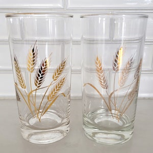 May include: Two clear drinking glasses with a gold wheat stalk design. The glasses have a gold rim and a simple, elegant shape. The wheat design is detailed and adds a touch of vintage charm to the glassware.