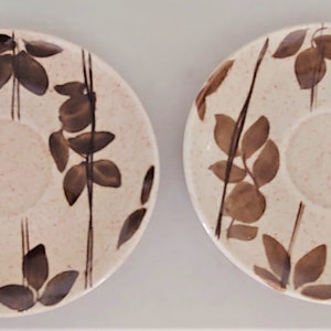 May include: Two white ceramic saucers with a brown leaf pattern. The saucers have a speckled glaze and a slightly raised rim.