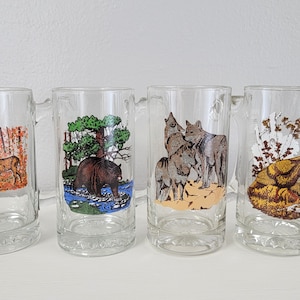 May include: Set of four clear glass mugs with handles. Each mug features a different nature scene with colorful illustrations. The mugs depict a deer, a bear, wolves, and a turkey.