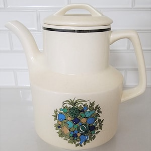 May include: A white ceramic coffee pot with a brown trim around the lid and a fruit design on the bottom. The design features a pineapple, pears, and other fruits in shades of green, blue, and orange.
