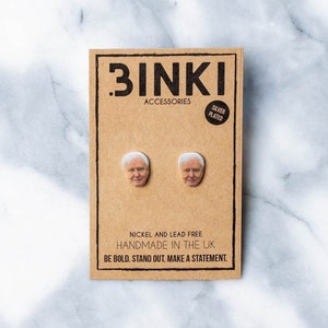Puede incluir: Pendientes de botón plateados con un diseño de rostro impreso, presentados en una tarjeta marrón. La tarjeta dice ".BINKI ACCESSORIES SILVER PLATED NICKEL AND LEAD FREE HANDMADE IN THE UK BE BOLD. STAND OUT. MAKE A STATEMENT."