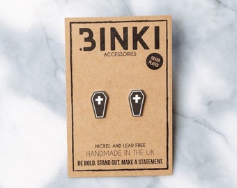 May include: A pair of silver plated coffin shaped earrings with a white cross on each. The earrings are handmade in the UK and are nickel and lead free. The packaging is brown cardboard with the text "BINKI ACCESSORIES SILVER PLATED" and "BE BOLD. STAND OUT. MAKE A STATEMENT."