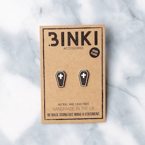 May include: A pair of silver plated coffin shaped earrings with a white cross on each. The earrings are handmade in the UK and are nickel and lead free. The packaging is brown cardboard with the text "BINKI ACCESSORIES SILVER PLATED" and "BE BOLD. STAND OUT. MAKE A STATEMENT."