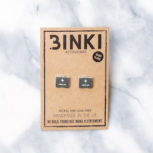 Könnte beinhalten: Ein Paar silberne Ohrringe in Form einer Erste-Hilfe-Box. Die Ohrringe befinden sich auf einer braunen Karte mit dem Text "BINKI ACCESSORIES SILVER PLATED" und "NICKEL AND LEAD FREE HANDMADE IN THE UK BE BOLD. STAND OUT. MAKE A STATEMENT."