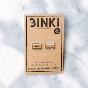 以下が含まれることがあります： 茶色のカードに銀メッキの カセットテープのイヤリングが2つ。カードには「BINKI ACCESSORIES SILVER PLATED」と「NICKEL AND LEAD FREE HANDMADE IN THE UK BE BOLD. STAND OUT. MAKE A STATEMENT.」の文字が書かれている
