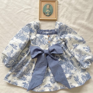 May include: A white and blue toile dress with a square neckline, long, puffy sleeves, and a large blue bow. The dress features a button-down front and a print of pastoral scenes. A framed portrait hangs above the dress.