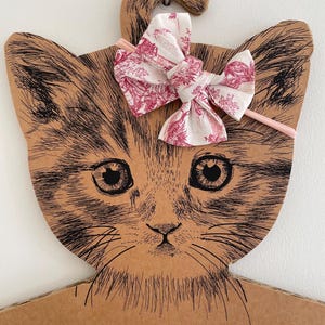 May include: A pink and white floral patterned headband with a bow. The headband is on a cardboard cat-shaped hanger.