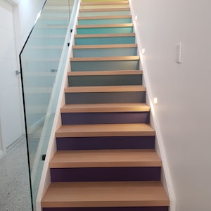 Puede incluir: Una escalera con peldaños de madera pintados en un degradado arcoíris, comenzando con verde y terminando con morado. La escalera tiene una barandilla de vidrio y paredes blancas.