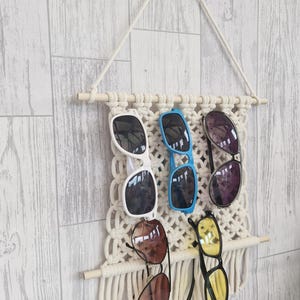 May include: A cream-coloured macrame wall hanging organiser featuring two horizontal wooden dowels and a braided cord for suspension. The woven cotton fibre grid holds five pairs of sunglasses in white, blue, black, brown, and yellow frames.