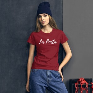 May include: A red t-shirt with the text "La Poete" in white lettering. The shirt is being worn by a person with a black beanie and blue jeans.