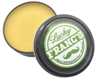 May include: Open tin of Lucky Franc's Heavy Hold Mustache Wax, unscented. The wax is a light yellow color and sits in a black tin with a green and white label. The label features the brand name and a mustache graphic.