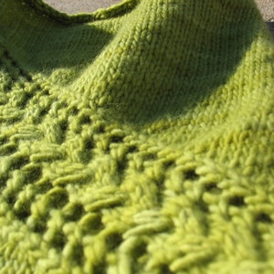 以下が含まれることがあります： 緑色の編み物のショールをクローズアップした写真です。ショールは柔らかな糸でできており、繊細で流れるようなデザインです。
