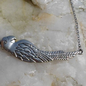 May include: A silver parrot pendant necklace. The pendant, with etched feather detailing and a bird's head, hangs from a fine chain. The necklace is shown against a light, textured background, showcasing the jewellery's design.