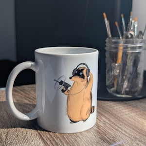 May include: White ceramic mug featuring a cartoon badger wearing headphones and holding a small device. The badger is tan and brown with a black mask. The mug has a curved handle and is set on a wooden surface.
