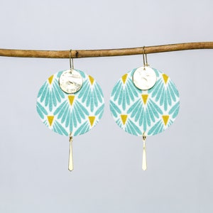 May include: A pair of turquoise and gold earrings with a geometric pattern. The earrings are round and have a small gold disc in the centre. The earrings are hanging from a gold hook.