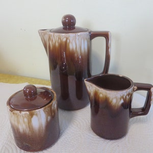 May include: A brown and white ceramic coffee pot, creamer, and sugar bowl set. The set has a drip glaze design.
