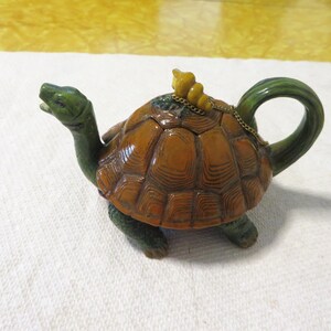 May include: A ceramic teapot in the shape of a turtle. The turtle has a brown shell, green head, legs, and handle. A small yellow figure sits on the shell, connected by a chain to the lid. The teapot is on a white surface.